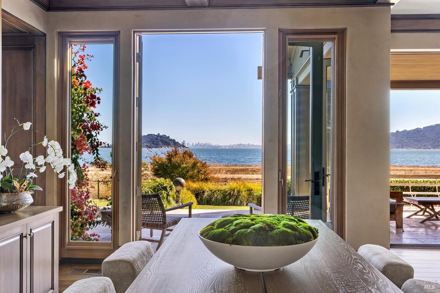This interior shot showcases a dining room with a stunning view of the water and distant cityscape through large, open doors and windows. A long wooden table is the centerpiece, adorned with a large bowl of moss, and the room is bathed in natural light, creating a serene and inviting atmosphere. The design emphasizes indoor-outdoor living, making it a desirable feature for potential buyers.