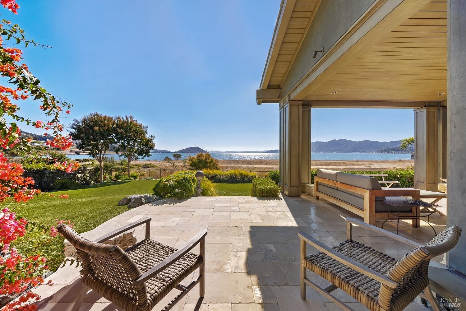 This image showcases a beautiful outdoor patio area with stunning waterfront views. The patio features stone flooring and comfortable wicker seating, creating an inviting space for relaxation and entertainment. The covered area provides shade, while the open view extends to the ocean, enhancing the property's appeal.