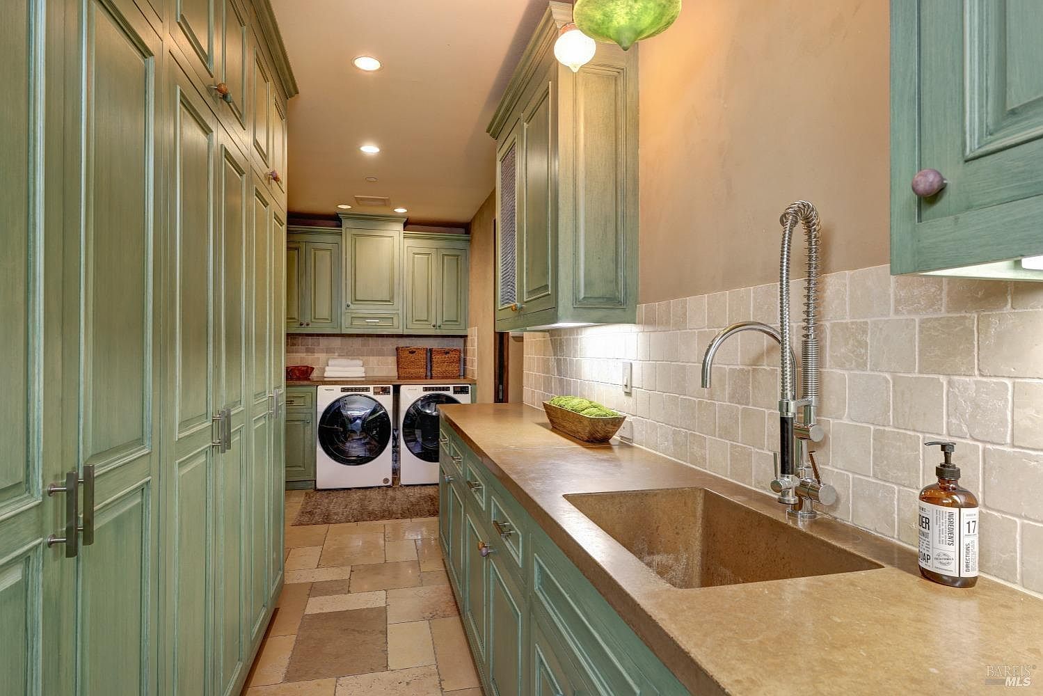 This is a well-appointed laundry room featuring custom green cabinetry with ample storage. A long countertop with a built-in sink provides workspace, and modern washing machines are neatly arranged. The room is illuminated by recessed lighting and pendant fixtures, creating a functional and stylish space.