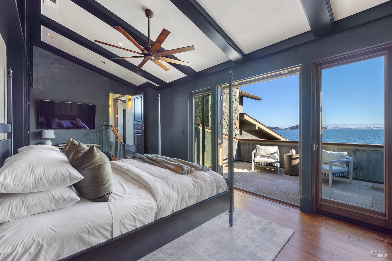 This is a primary bedroom featuring a four-poster bed with white linens and decorative pillows. The room has dark walls and exposed ceiling beams, with a ceiling fan overhead. A large sliding glass door opens onto a balcony with ocean views, creating a luxurious and serene atmosphere.