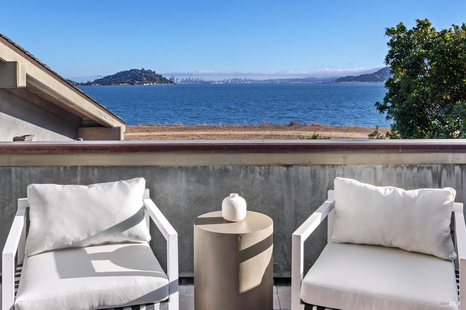 This image showcases a serene outdoor patio or balcony setting with a stunning waterfront view. Two white armchairs with cushions flank a cylindrical side table topped with a decorative vase. The scene evokes a sense of relaxation and luxury, highlighting the property's desirable outdoor living space and scenic location.