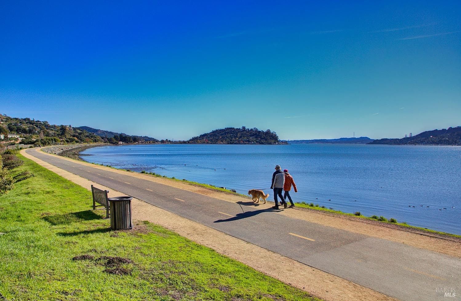 This image showcases a scenic walking path alongside a tranquil body of water, likely part of a community amenity. The path is well-maintained, with a bench and trash receptacle visible, suggesting a clean and inviting environment. Two people are walking a dog on the path, emphasizing the recreational opportunities available.