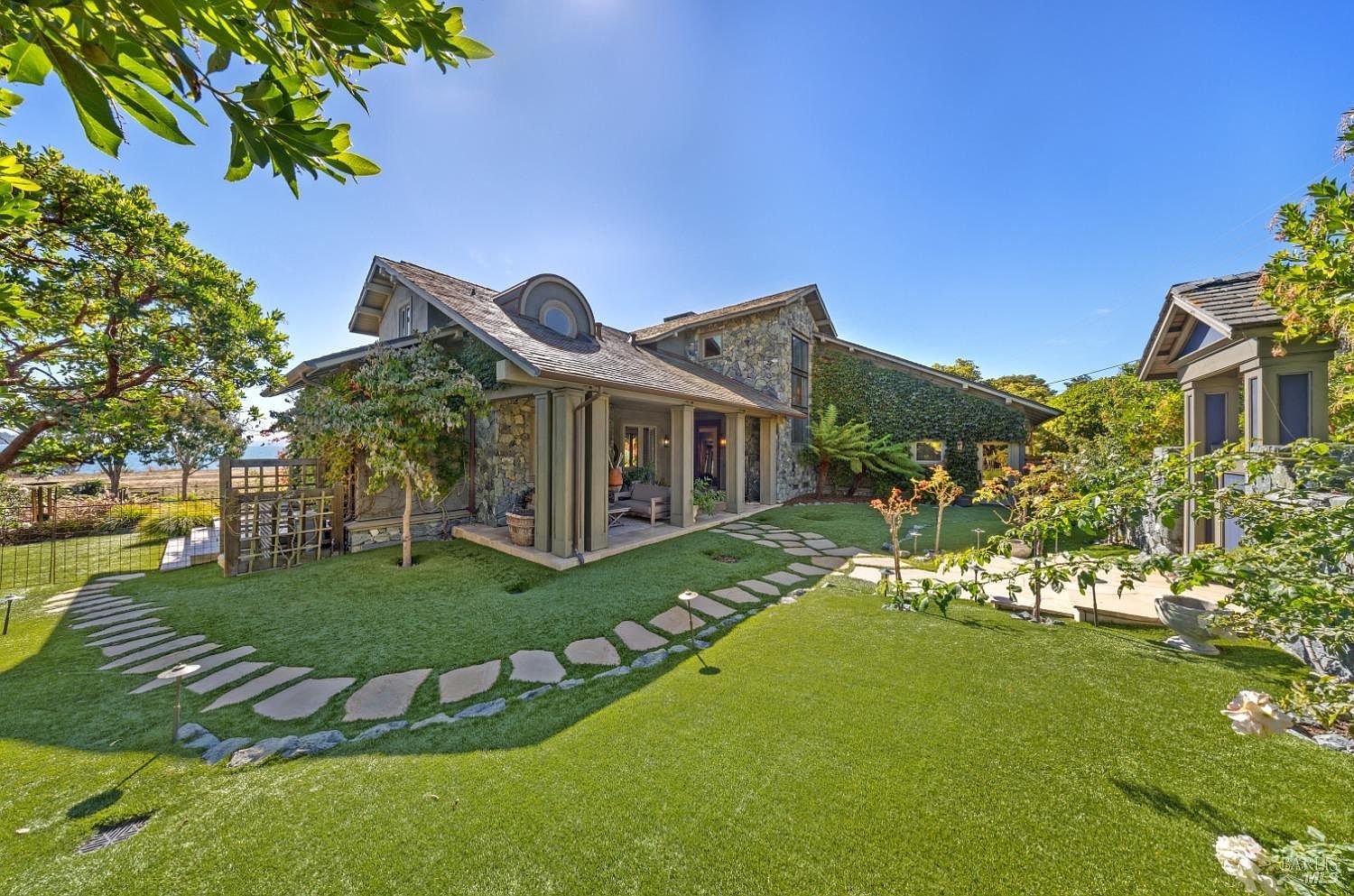 This image showcases a beautifully landscaped yard with a stone pathway leading to a charming stone house. The lush green lawn and mature trees create a serene and inviting atmosphere. The house features stone accents and a covered porch, blending seamlessly with the natural surroundings.