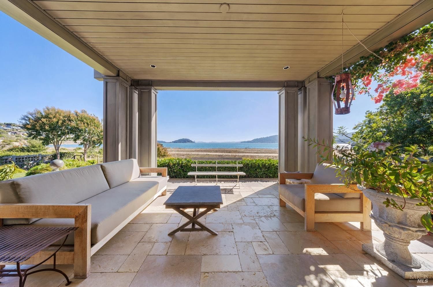 This image showcases a luxurious outdoor patio or balcony area with a stunning waterfront view. The space is furnished with comfortable seating, including a sofa and armchair, arranged around a coffee table. The architectural design features sturdy columns and a covered ceiling, providing shade and shelter, while the open design allows for unobstructed views of the water and distant landscape.