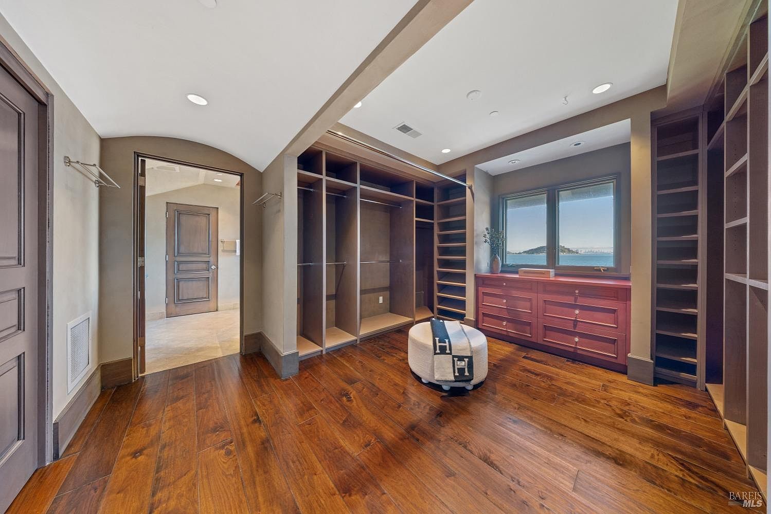 This is a spacious walk-in closet featuring hardwood floors, custom shelving, and a window with a view. The closet includes built-in drawers and ample hanging space, providing plenty of storage. The overall impression is luxurious and well-organized.