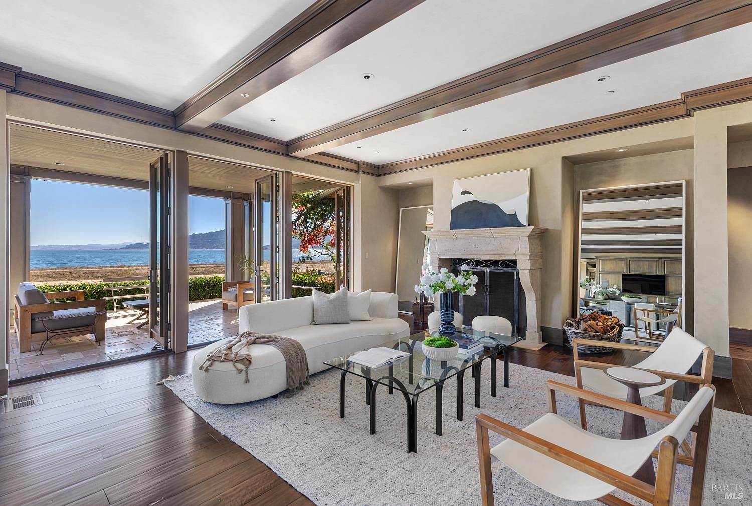 This is an interior shot of a luxurious living room with a stunning ocean view. The room features a white sofa, a glass coffee table, and two armchairs, all arranged on a light-colored rug. Large folding doors open to a patio, seamlessly blending indoor and outdoor living, while a fireplace and elegant decor add to the room's sophisticated ambiance.