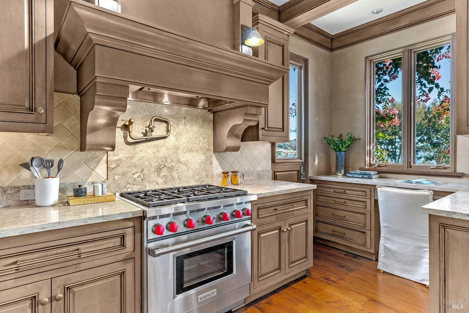 This kitchen features custom cabinetry in a warm, neutral tone, complemented by light countertops and a marble backsplash. A professional-grade stainless steel range with red knobs is the focal point, situated beneath a substantial range hood. Natural light streams in through a window, enhancing the inviting atmosphere of the space.