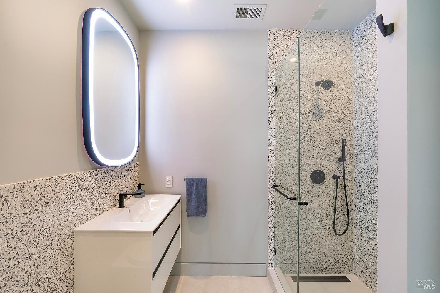 This modern bathroom features a sleek floating vanity with a white basin and a striking, oval-shaped LED-backlit mirror. The space is accented by a walk-in shower enclosed in glass, showcasing a contemporary terrazzo-style tile pattern that extends along the wall. The overall aesthetic is minimalist and sophisticated, emphasizing clean lines and high-end finishes.