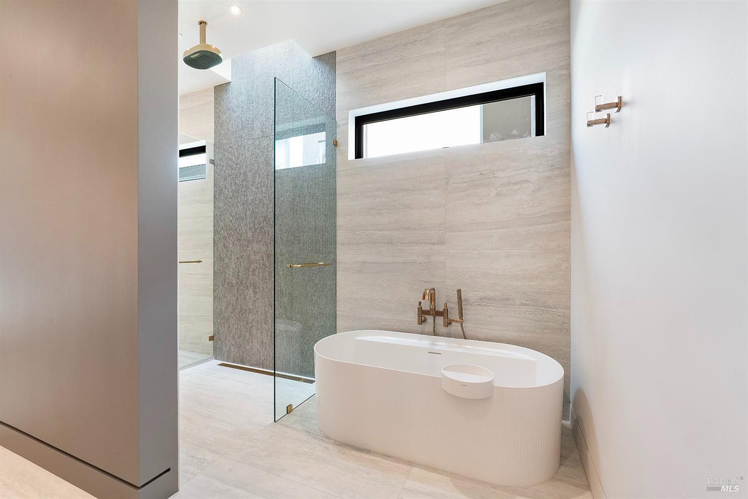 This luxurious primary bathroom features a sleek, modern design with a freestanding white soaking tub and a spacious walk-in shower enclosed by a glass panel. The room is accented by warm-toned, textured wall tiles and elegant gold-finished fixtures, creating a spa-like atmosphere. A horizontal window above the tub allows for natural light, enhancing the clean and sophisticated aesthetic of the space.