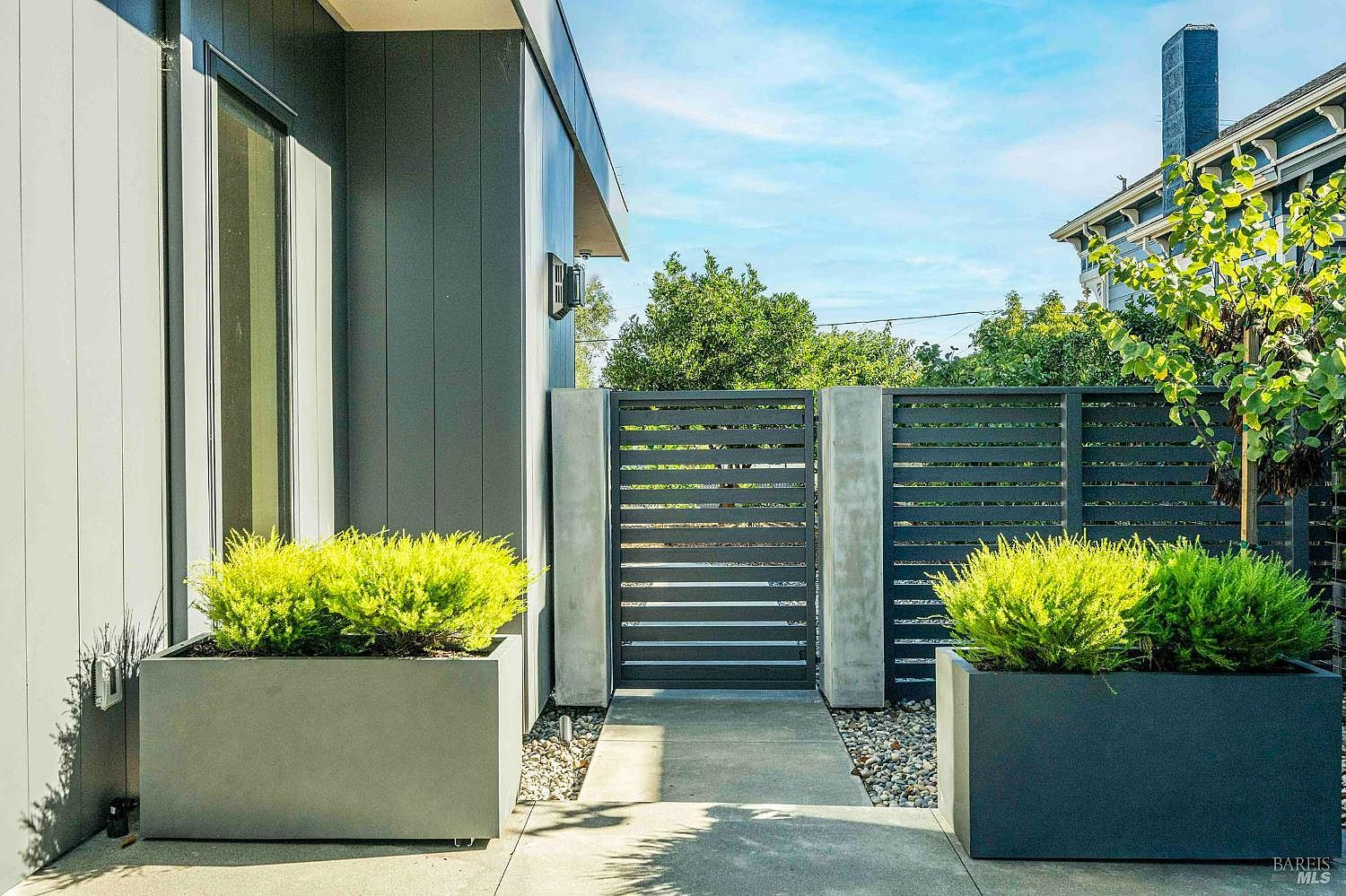 This modern entryway features a sleek, minimalist design with a concrete walkway leading to a contemporary horizontal-slat metal gate. Large, rectangular grey planters filled with vibrant green shrubs frame the path, creating a sophisticated and welcoming aesthetic. The clean lines and neutral color palette emphasize a high-end, architectural style typical of modern residential design.