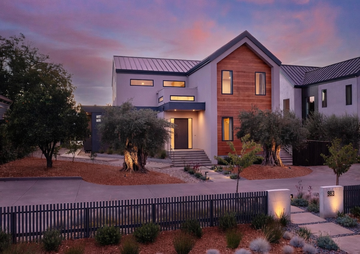This stunning modern residence features a striking blend of gray stucco and warm wood siding, accented by a sleek metal roof. The front facade is beautifully illuminated at dusk, highlighting the mature olive trees and the clean, geometric lines of the entryway. A contemporary fence and manicured landscaping frame the property, creating an inviting and sophisticated curb appeal.