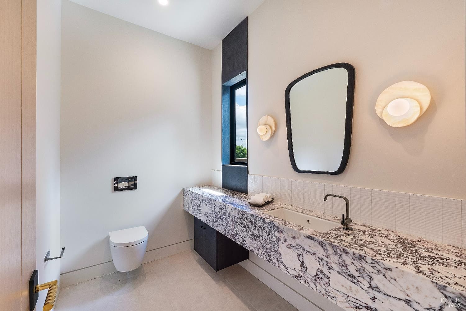 This modern powder room features a striking, thick-profile marble vanity with a sleek, integrated sink and dark faucet fixtures. The space is accented by a minimalist black-framed mirror, unique circular wall sconces, and a tall, narrow window set against a dark, textured accent wall. The overall aesthetic is sophisticated and contemporary, utilizing high-contrast materials to create a bold, luxurious atmosphere.