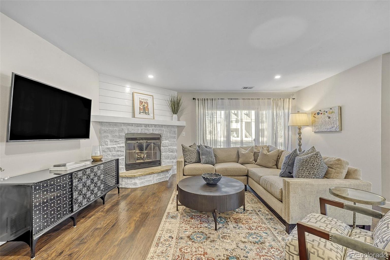 This is a cozy living room featuring a large sectional sofa, a round coffee table on a patterned rug, and a fireplace with a TV mounted above a modern cabinet. The room is well-lit with natural light from the windows and complemented by a floor lamp, creating a warm and inviting atmosphere. The overall style is contemporary with a touch of traditional elements.