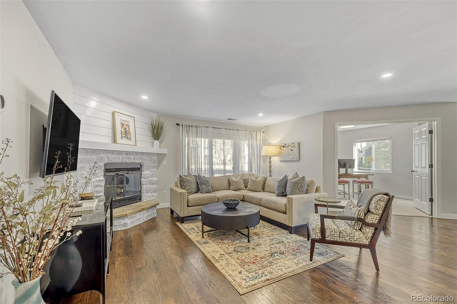 This is an interior shot of a living room featuring a sectional sofa, a round coffee table on a patterned rug, and an accent chair. A fireplace with a white mantel and a mounted TV are visible on one wall, while sheer curtains adorn the windows. The room has hardwood floors and an open doorway leading to another room, creating a warm and inviting atmosphere.