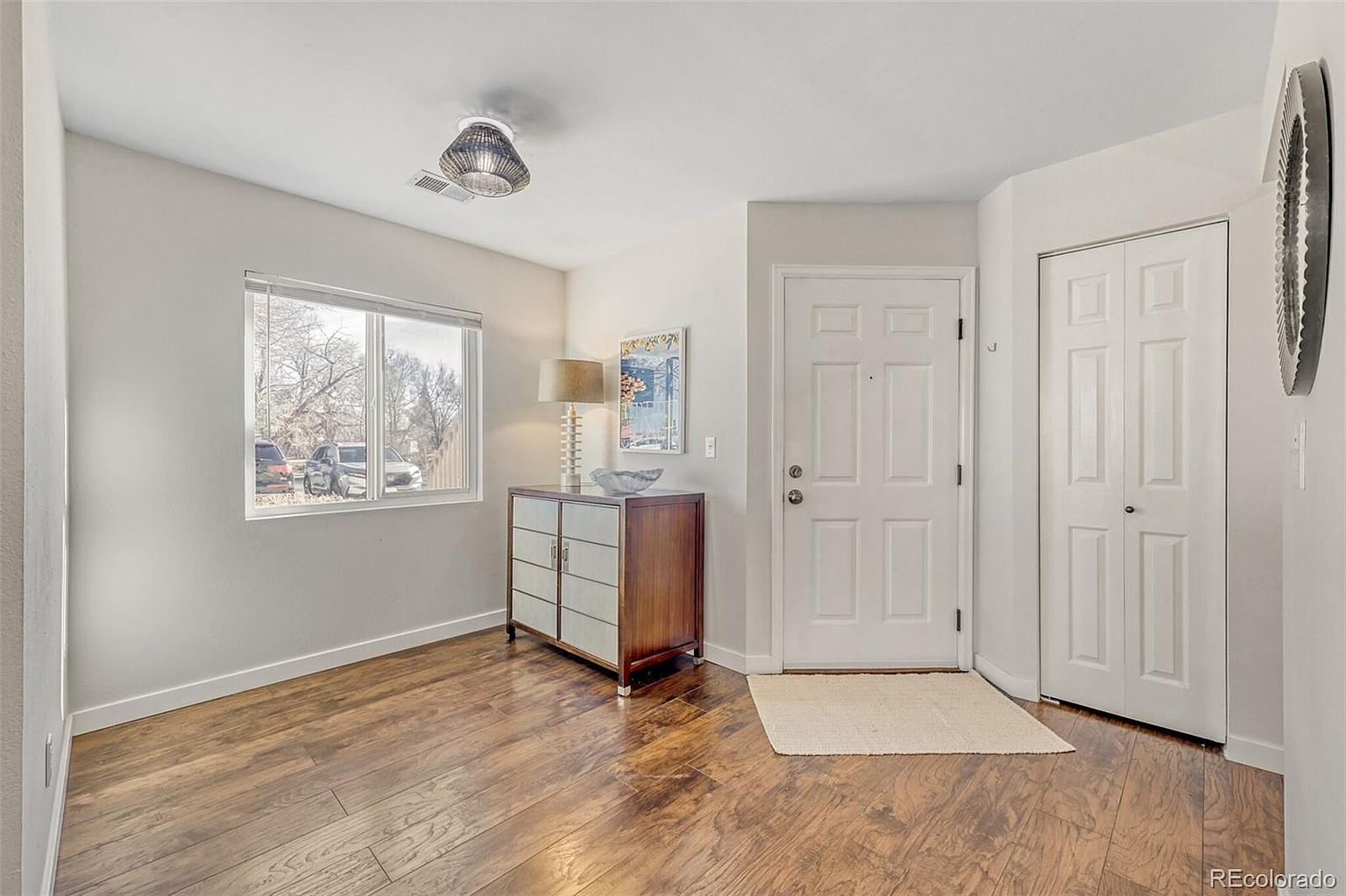 This is an interior shot of a home's entryway. The space features hardwood flooring, a window providing natural light, and a decorative cabinet with a lamp. A white front door and a closet door are visible, along with a neutral-colored rug, creating a welcoming and tidy first impression.