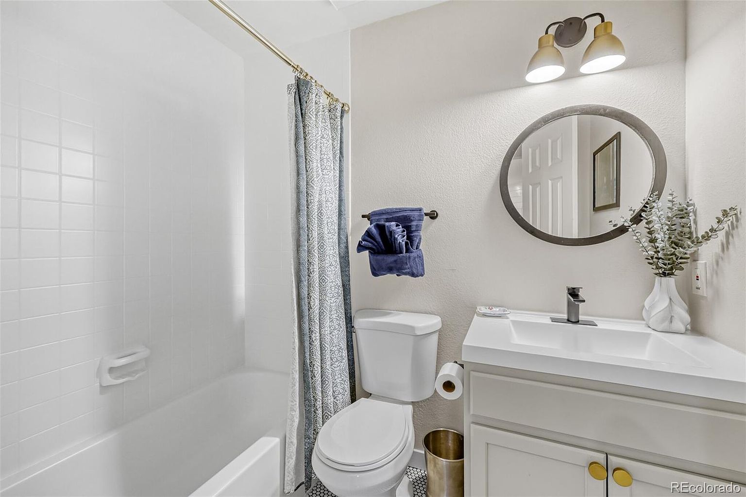 This is a well-lit bathroom featuring a bathtub with white tile surround and a patterned shower curtain. The room includes a toilet, a vanity with a white countertop and gray cabinets, and a round mirror above the sink. The overall impression is clean and functional, suitable for a guest or secondary bathroom.