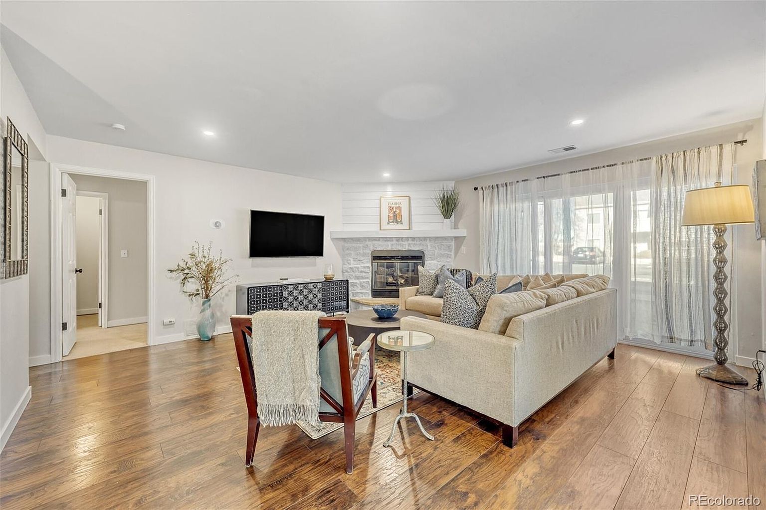 This is a well-lit living room featuring hardwood floors and a neutral color palette. A comfortable beige sofa is positioned near a fireplace with a white mantel, and a flat-screen TV is mounted on the wall. The room is decorated with stylish furniture and accessories, creating a cozy and inviting atmosphere.