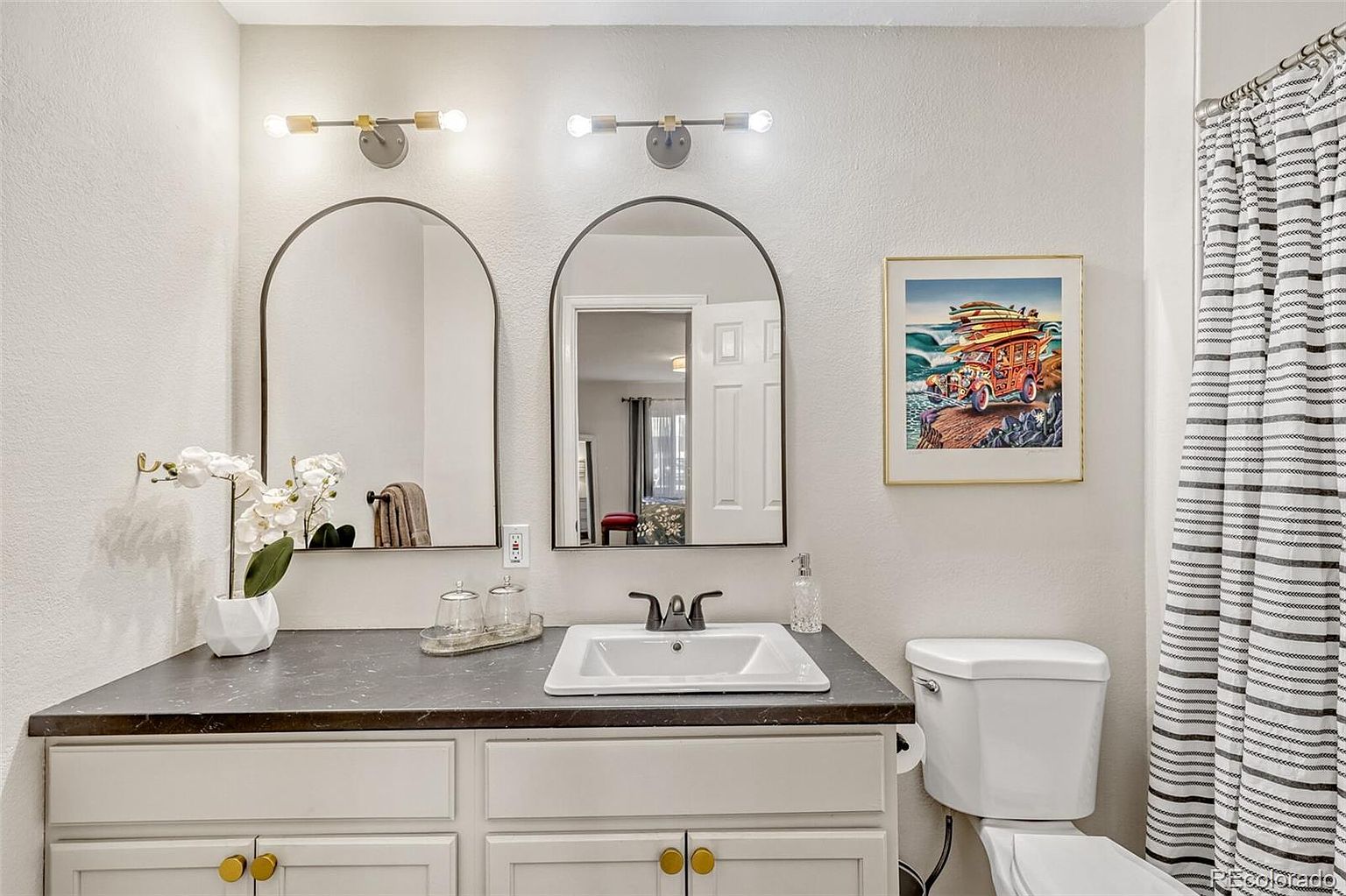 This is a well-lit bathroom featuring a vanity with a dark countertop and white cabinets with gold knobs. Above the vanity are two arched mirrors with modern light fixtures. A framed art piece hangs above the toilet, and a striped shower curtain adds a touch of style to the space.