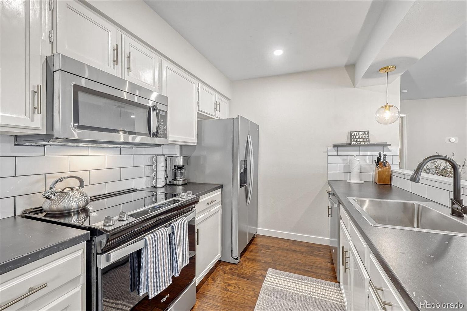 This is a well-lit kitchen featuring white cabinets, stainless steel appliances, and dark countertops. The kitchen includes a microwave, oven, refrigerator, and a sink with a modern faucet. The flooring is dark wood, and a small rug is placed near the sink, creating a cozy and functional space.