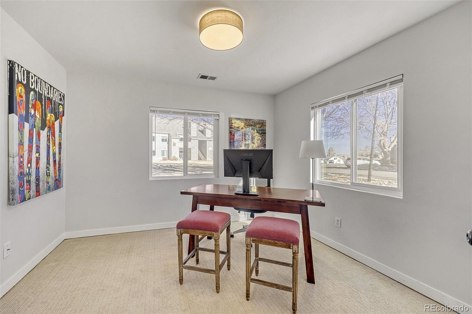 This is an interior shot of a home office featuring a wooden desk with a computer monitor, two red cushioned stools, and artwork on the walls. Natural light streams in through two windows, creating a bright and inviting workspace. The room has a neutral color palette with beige carpet and white walls, contributing to a clean and modern aesthetic.