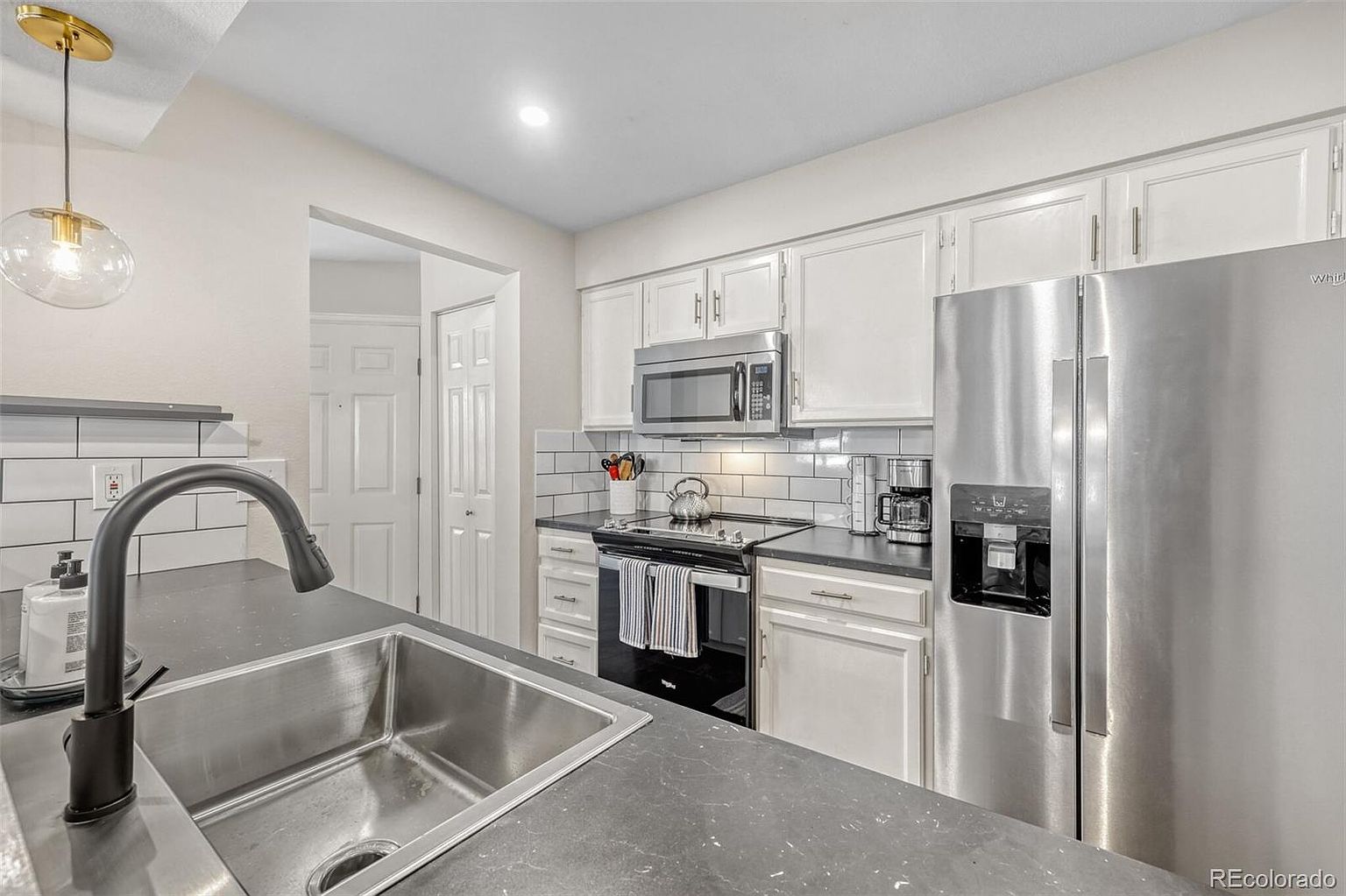 This is a well-lit kitchen featuring white cabinets, stainless steel appliances, and dark countertops. The kitchen includes a stainless steel sink with a black faucet, a microwave, an oven, and a refrigerator. The backsplash is white subway tile, and the overall impression is clean and modern.