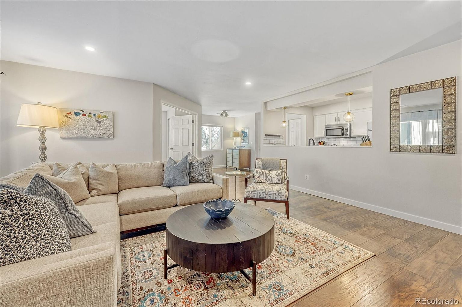 This is an inviting living room featuring a comfortable sectional sofa adorned with throw pillows, a round wooden coffee table, and a patterned area rug. The room has hardwood flooring and neutral-colored walls, complemented by a decorative mirror and artwork. An open layout connects the living room to the kitchen, creating a seamless flow.