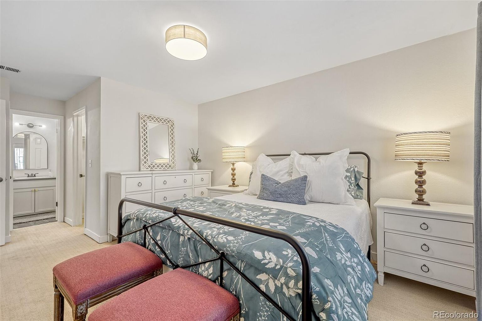 This is a cozy bedroom featuring a metal-framed bed with a blue floral comforter and decorative pillows. The room is furnished with a white dresser, a matching nightstand, and two upholstered ottomans at the foot of the bed. A bathroom entrance is visible, suggesting an en-suite arrangement, and the overall aesthetic is clean and inviting.