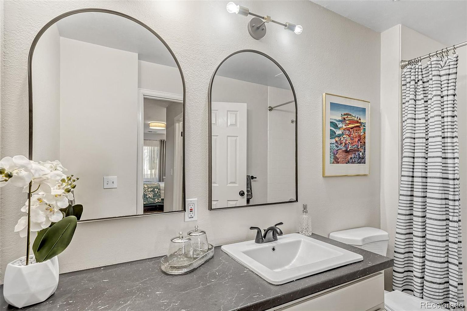 This is a well-lit bathroom featuring a double vanity with a dark countertop and a white sink. Two arched mirrors hang above the vanity, reflecting the room and adding a touch of modern style. A striped shower curtain is visible, and a framed artwork hangs on the wall, contributing to the room's aesthetic appeal.