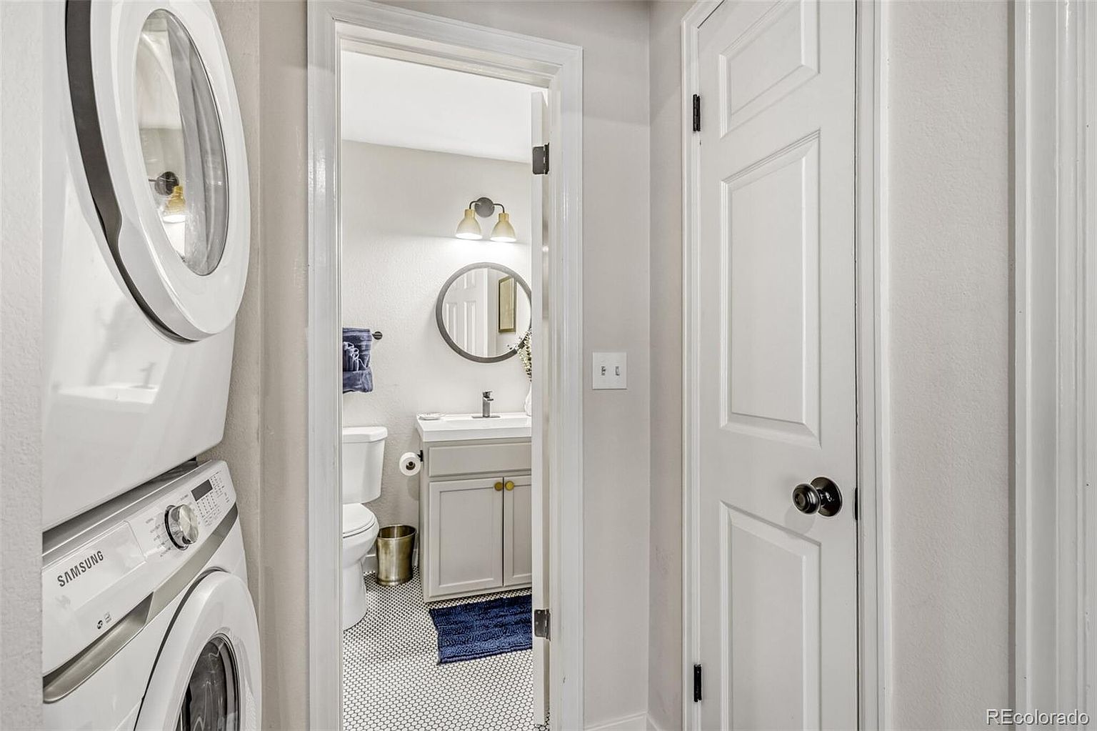 The image showcases a laundry area adjacent to a bathroom. A stacked washer and dryer unit is visible on the left, while an open doorway reveals a bathroom with a toilet, vanity, round mirror, and black and white tiled floor. The color palette is neutral, with white and gray tones dominating the scene, creating a clean and functional space.