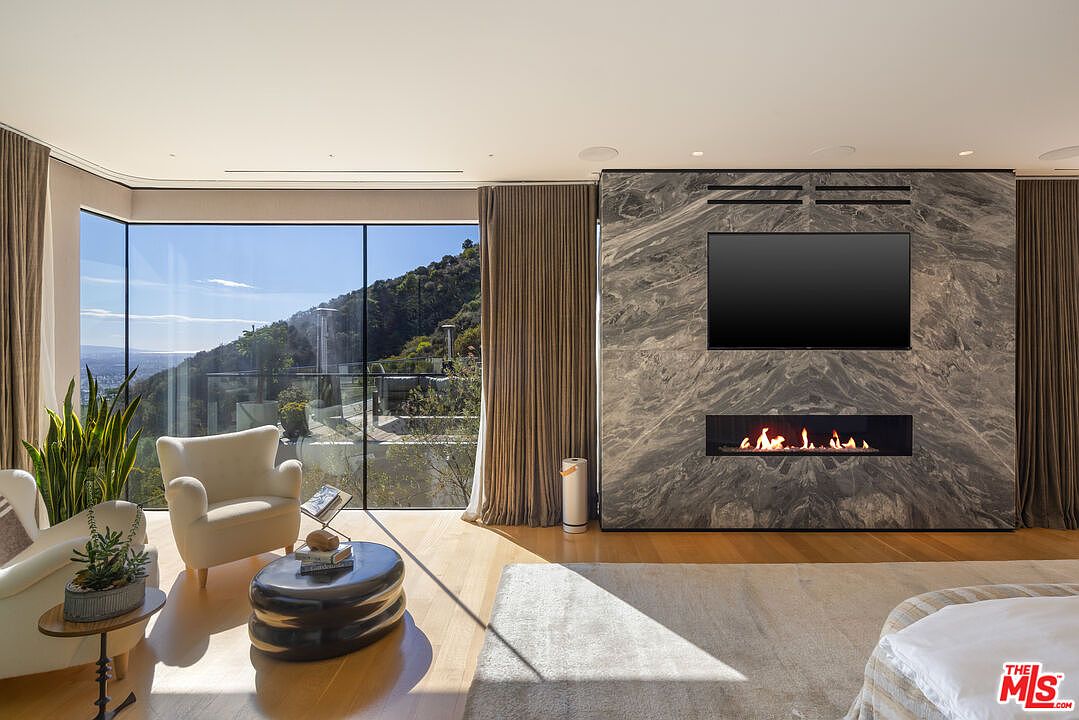 This spacious primary bedroom features a striking stone-clad chimney breast that serves as a focal point, housing a television and a linear fireplace. Large, expansive windows offer panoramic views of the surrounding hills, while light wood flooring and neutral furnishings create a serene, modern atmosphere. The room is bathed in natural light, emphasizing the clean lines and sophisticated design of the space.