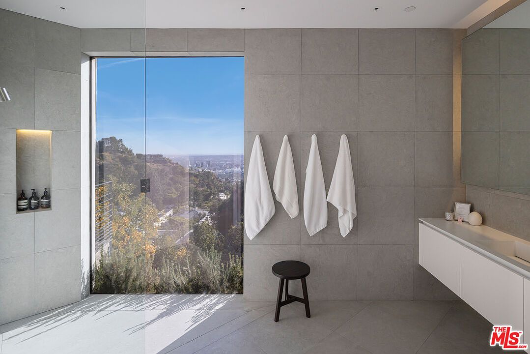 This modern primary bathroom features large-format gray stone wall tiles and a minimalist white floating vanity. A floor-to-ceiling glass panel separates the shower area, which offers a stunning, unobstructed view of the surrounding hills and city skyline. A simple black stool sits against the wall, complemented by four white towels hanging neatly, creating a serene and high-end spa-like atmosphere.