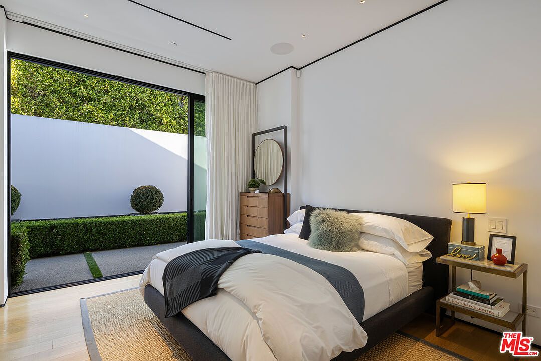 This modern bedroom features a neutral color palette with a dark upholstered bed frame and a textured rug. A large sliding glass door provides a seamless transition to an outdoor patio area with manicured hedges and a white privacy wall. The room is accented by a wooden dresser with a circular mirror and a bedside table with a contemporary lamp, creating a serene and minimalist atmosphere.