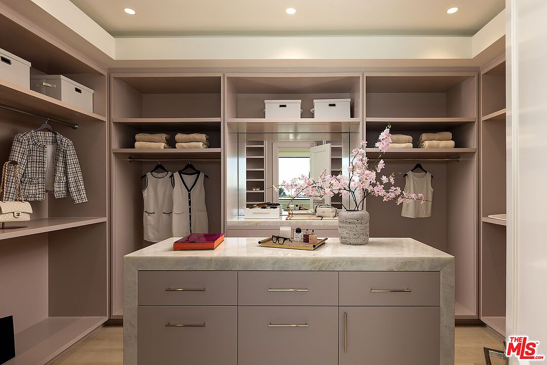 This elegant walk-in closet features custom built-in cabinetry in a soft, neutral tone with integrated hanging rods and open shelving. A central island with a stone countertop provides a sophisticated focal point, topped with a decorative vase of cherry blossoms and personal accessories. The space is illuminated by recessed lighting, creating a bright and organized atmosphere suitable for a luxury home.
