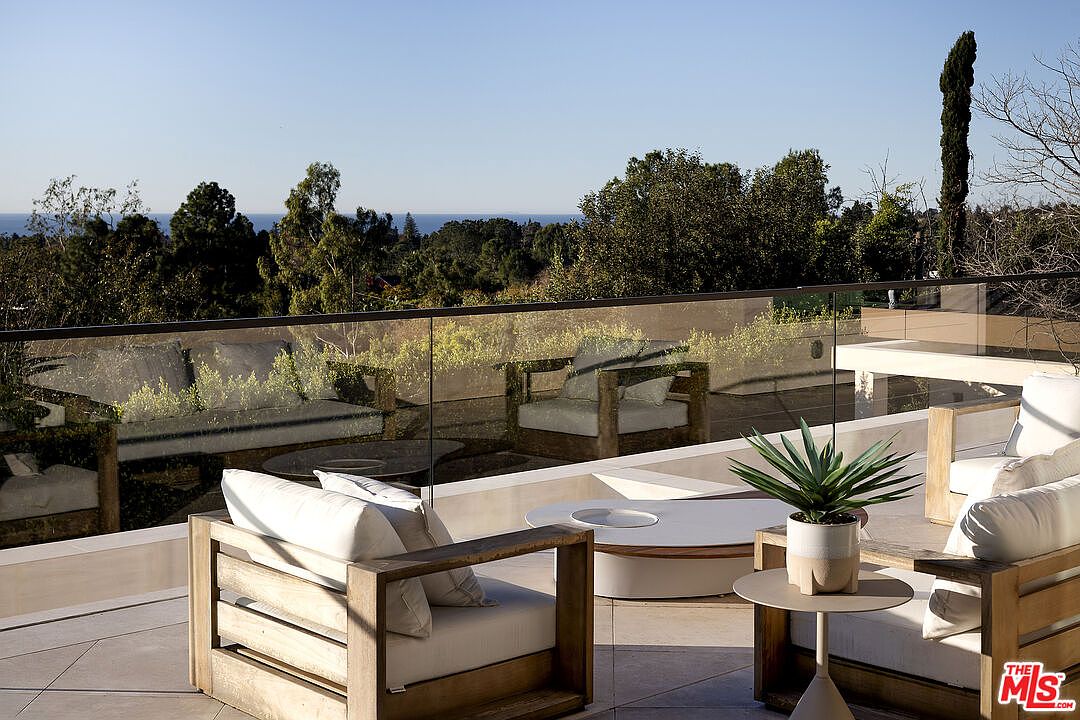 This expansive outdoor terrace features a modern aesthetic with light-toned stone flooring and a sleek glass railing that provides an unobstructed view of the surrounding trees and distant ocean. The space is furnished with comfortable, light-wood framed seating arranged around a low, circular coffee table, creating an inviting lounge area. The scene is captured from a slightly elevated perspective, emphasizing the seamless transition between the luxury living space and the natural landscape.