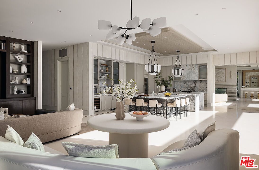 This expansive, open-concept living and kitchen area features a sophisticated neutral palette and high-end finishes. A large central island with stone countertops and bar seating serves as the focal point, complemented by a round dining table in the foreground and a built-in shelving unit to the left. The space is illuminated by a striking modern chandelier and dual pendant lights, creating an airy and luxurious atmosphere suitable for entertaining.