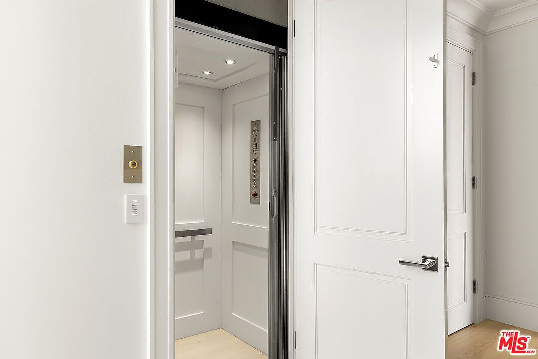 This image captures a private residential elevator with an open door, revealing a clean, minimalist interior with recessed lighting and a vertical control panel. To the right, a white paneled door with a modern lever handle leads into an adjacent room, while a wall-mounted elevator call button and a small control interface are visible on the left. The space features light-toned flooring and a bright, neutral color palette, creating a sophisticated and functional transition area.