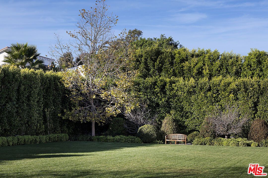 This expansive, well-manicured lawn serves as a private backyard retreat, framed by dense, mature hedges that provide significant privacy. A wooden bench sits nestled near the greenery, offering a quiet spot to enjoy the landscape, while a prominent deciduous tree stands as a focal point in the center of the yard. The perspective is a wide, eye-level shot that captures the lush greenery and open space, suggesting a serene and secluded outdoor environment.