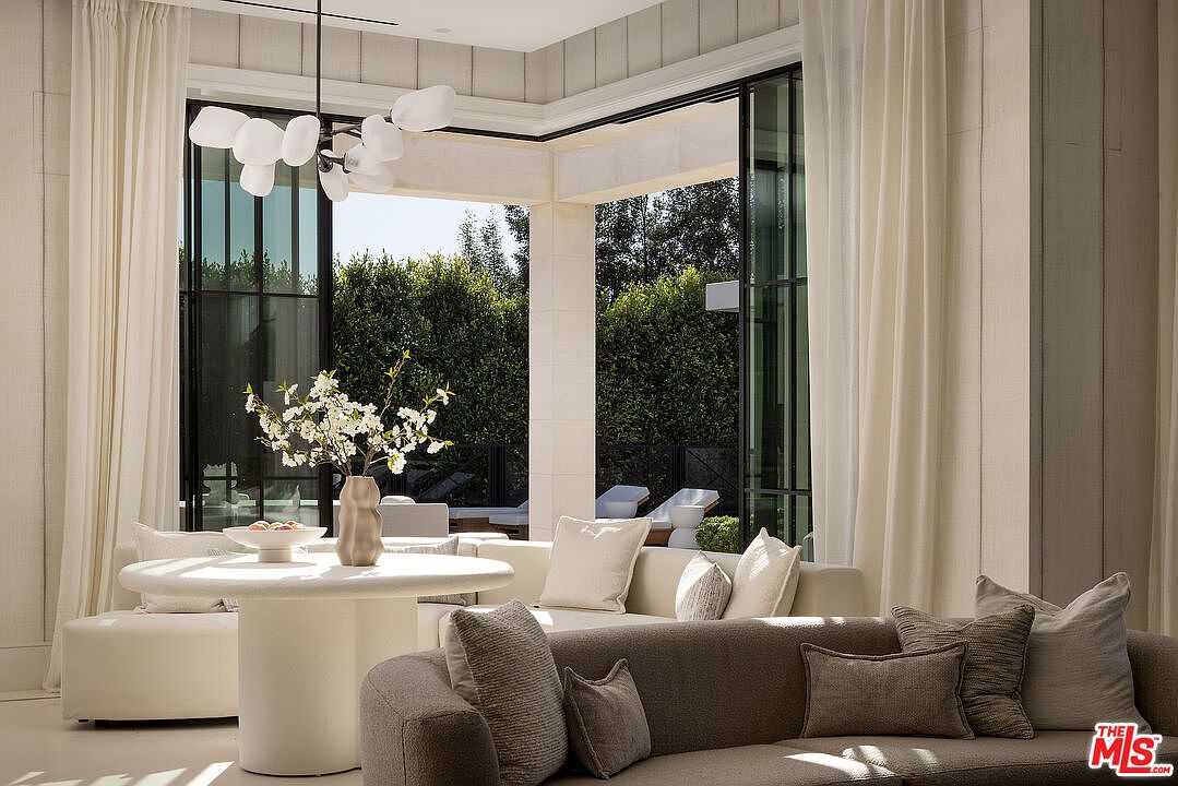 This elegant living area features a sophisticated neutral color palette and a seamless transition to the outdoors through large glass doors. The space is anchored by a curved sofa and a sculptural round table, complemented by a unique, organic-shaped pendant light fixture. The bright, airy atmosphere is enhanced by soft drapery and a view of lush greenery, creating a serene and luxurious indoor-outdoor living experience.
