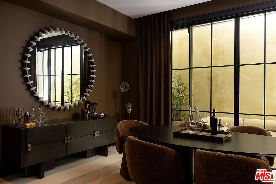 This sophisticated dining room features a dark, moody aesthetic with a large oval table surrounded by textured, upholstered chairs. A prominent circular mirror with a decorative, multi-faceted frame hangs above a sleek, dark sideboard, creating a focal point against the deep-toned walls. Large, grid-paned windows allow natural light to filter in from an adjacent outdoor space, enhancing the room's elegant and intimate atmosphere.