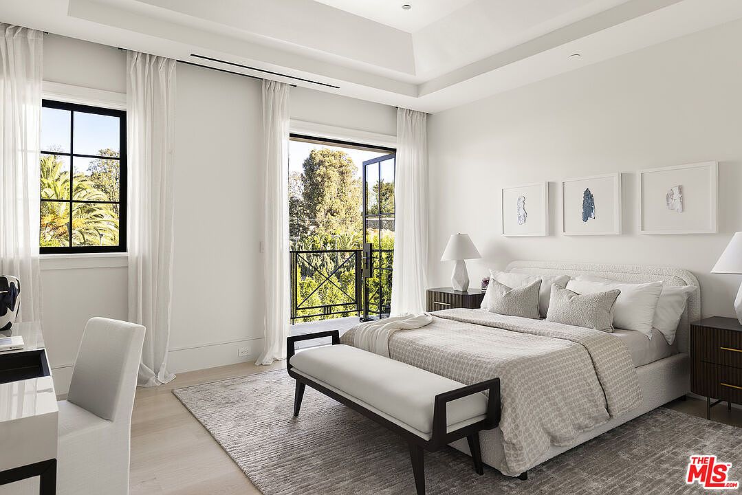 This bright and airy primary bedroom features a neutral color palette, a tray ceiling, and light wood flooring. A plush bed with a textured headboard is centered in the room, flanked by two dark wood nightstands and topped with minimalist artwork. Large windows and a glass door leading to a private balcony fill the space with natural light, creating a serene and sophisticated atmosphere.