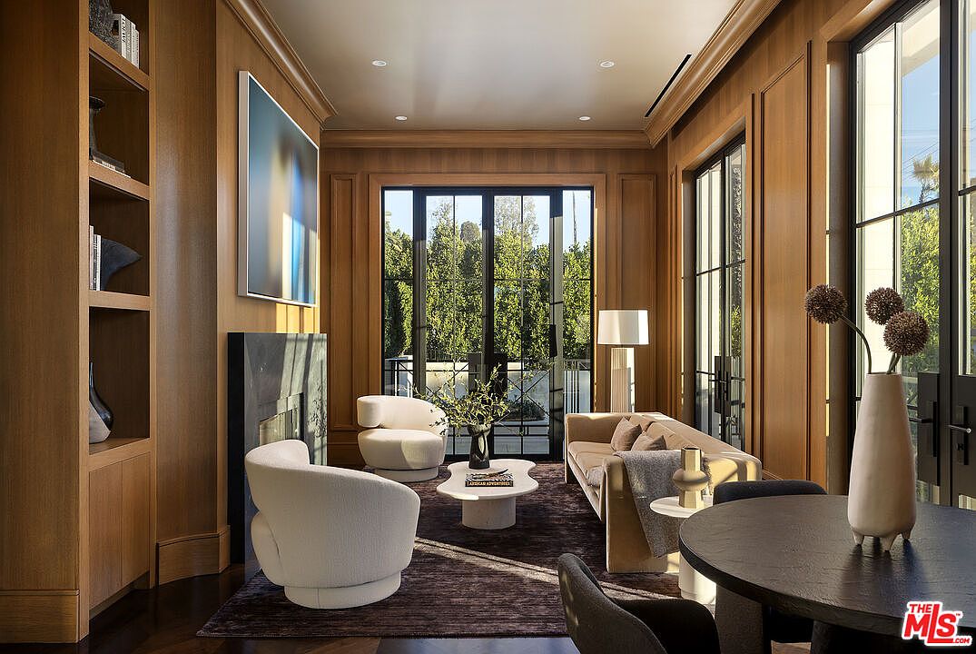 This sophisticated living room features warm wood-paneled walls and a dark stone fireplace surround, creating a cozy yet refined atmosphere. The space is furnished with plush, cream-colored seating, a central coffee table, and a sofa, all arranged on a dark area rug. Large, black-framed glass doors provide ample natural light and a view of the lush greenery outside, while a circular dining table in the foreground adds versatility to the room's layout.