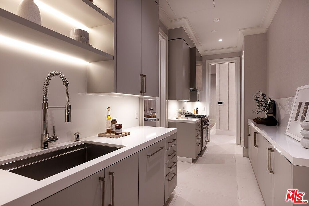 This sleek, modern kitchen features a galley-style layout with light gray cabinetry and crisp white countertops. A professional-style faucet and deep sink are positioned under integrated under-cabinet lighting, while a secondary cooking area is visible in the background. The minimalist aesthetic is enhanced by clean lines, recessed lighting, and a neutral color palette that creates a bright and sophisticated atmosphere.