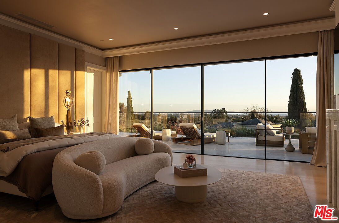This luxurious primary bedroom features a neutral color palette with textured wall paneling behind the bed and a curved, cream-colored sofa positioned in the center of the room. Large, floor-to-ceiling glass sliding doors open onto a spacious terrace, providing an expansive view of the surrounding landscape. The room is bathed in warm, golden-hour light, creating a serene and sophisticated atmosphere.