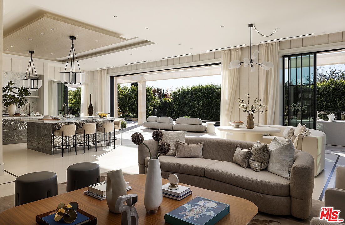 This expansive open-concept living space seamlessly blends a sophisticated lounge area with a gourmet kitchen and dining nook. The room features a neutral color palette, plush curved seating, and large glass doors that slide open to reveal a lush outdoor garden. The design emphasizes a luxurious, airy atmosphere with high-end finishes, designer lighting fixtures, and a curated selection of decor on the foreground coffee table.