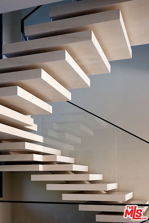 This image captures a striking, modern floating staircase featuring thick, light-toned stone treads that appear to hover without visible supports. A sleek, transparent glass railing runs alongside the stairs, providing a clean and minimalist aesthetic. The perspective is angled from below, emphasizing the geometric precision and architectural elegance of the design.