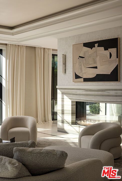 This sophisticated living area features a contemporary aesthetic with a neutral color palette and soft, rounded furniture silhouettes. A prominent stone-clad chimney breast serves as the focal point, housing a modern fireplace, while an abstract piece of art hangs above. The space is framed by tall, light-colored drapery and includes plush, textured seating that creates an inviting and luxurious atmosphere.