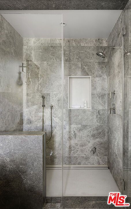 This modern walk-in shower features large-format grey stone tiles on the walls and a matching stone-clad partition wall. The space includes a wall-mounted shower head, a handheld sprayer, and a recessed niche for storage, all enclosed by a clear glass panel. The minimalist design is complemented by a clean, light-colored shower floor and polished metal fixtures.