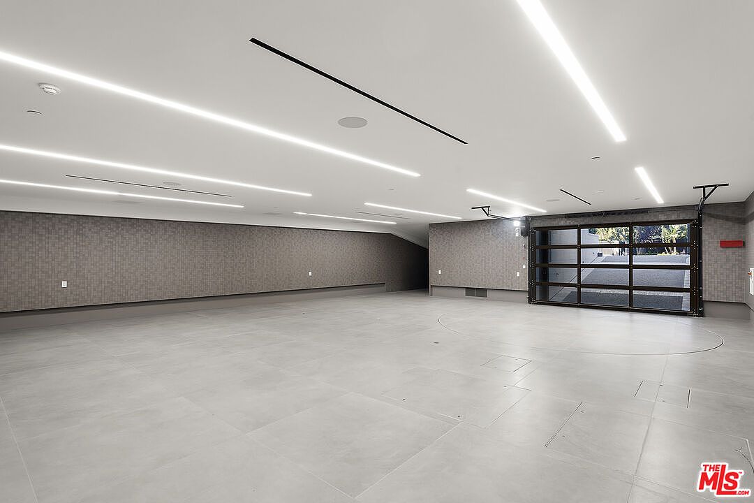This expansive, modern garage features a minimalist aesthetic with large-format light gray floor tiles and textured, dark-toned wall paneling. A prominent circular turntable is integrated into the floor, designed for easy vehicle maneuvering, while a multi-pane glass garage door provides a view of the driveway outside. The ceiling is finished with recessed linear lighting strips, creating a sleek and high-end atmosphere suitable for a luxury car collection.