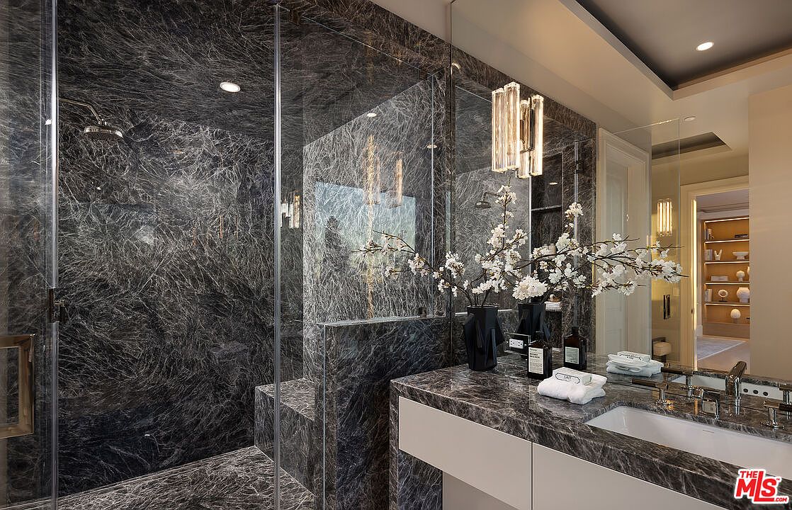 This luxurious bathroom features dark, heavily veined stone surfaces throughout the shower enclosure and vanity area. A glass-enclosed walk-in shower with a built-in bench sits adjacent to a sleek vanity, accented by modern, multi-tiered glass pendant lighting. The perspective captures the sophisticated, high-contrast aesthetic, looking from the vanity toward the shower and into an adjacent hallway.