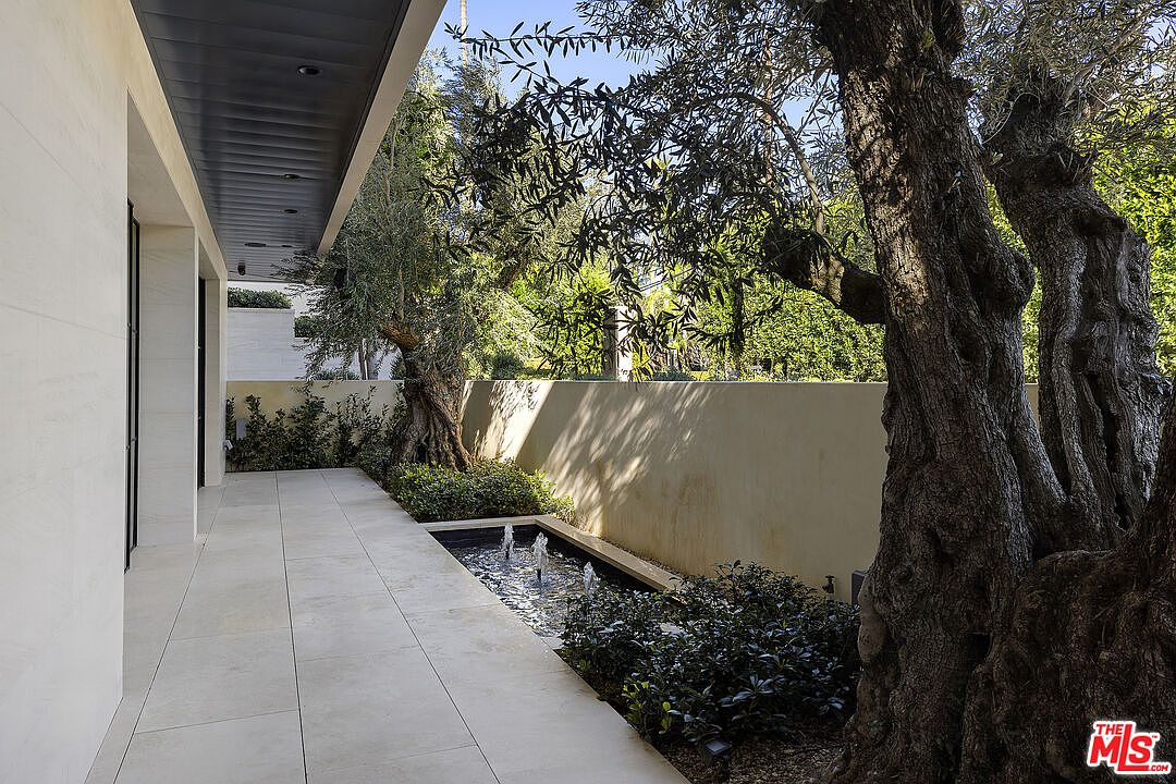 This serene outdoor patio features large, light-toned stone pavers and a sleek, modern water feature with multiple fountain jets. The space is framed by a tall, smooth wall and mature olive trees, creating a private and tranquil atmosphere. The perspective looks along the covered walkway, highlighting the clean architectural lines and the integration of nature with the built environment.